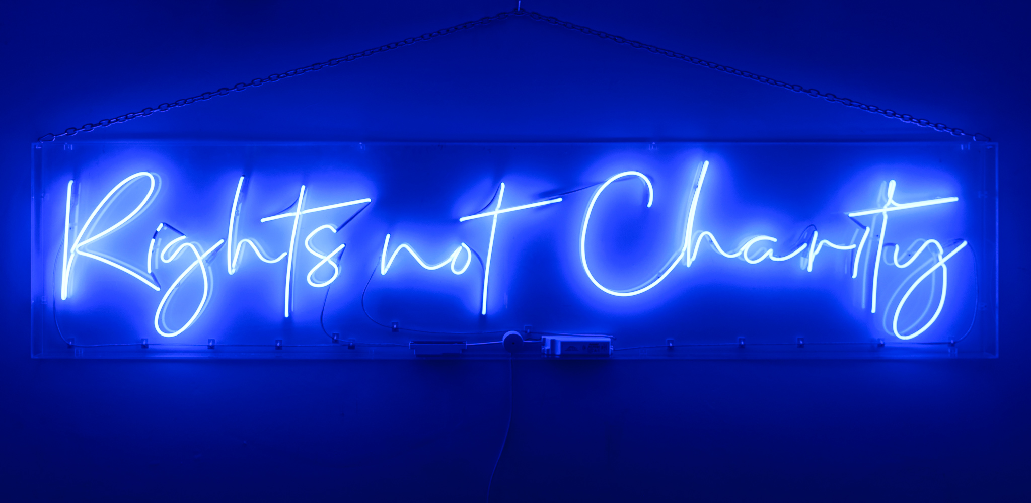 The wording reads on the artwork. 'Rights not Charity'- blue marine neon lights in a stylised handwritten font. The neon words are framed inside a transparent perspex box.