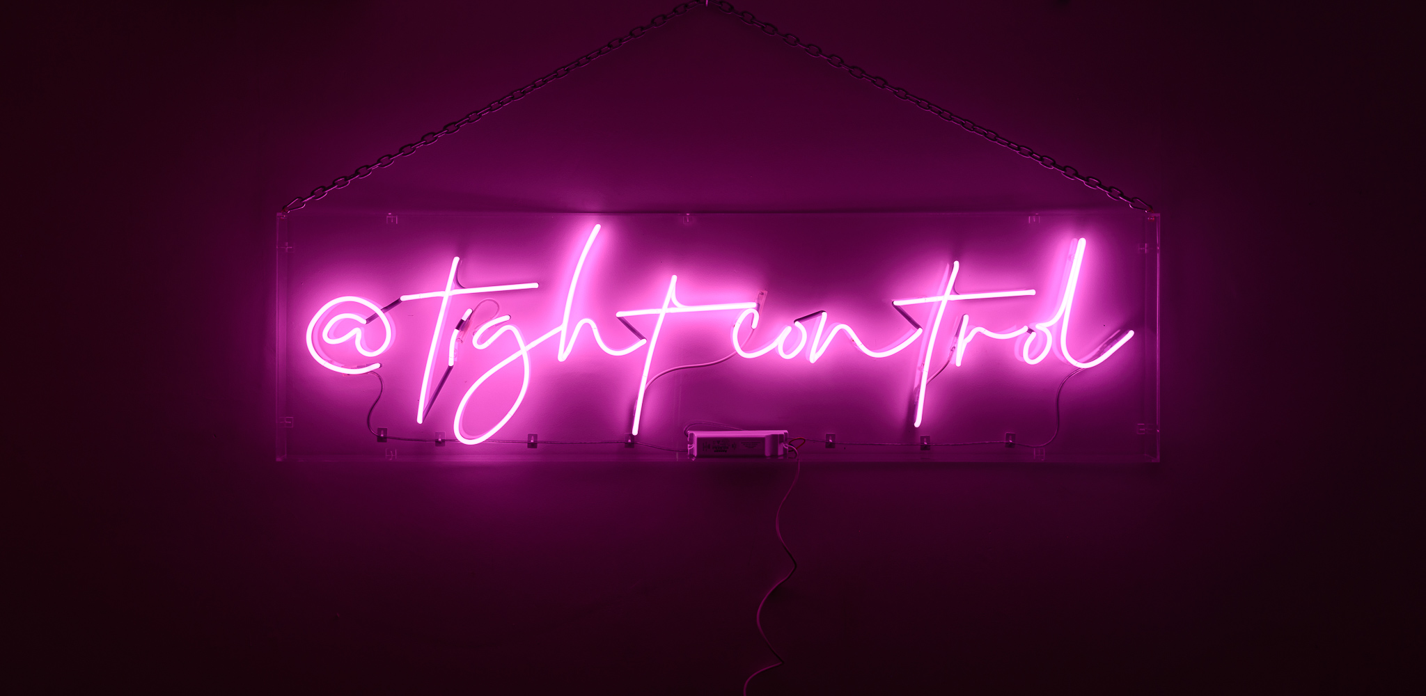 The wording reads on the artwork '@TightControl'- neon lights in shocking pink lights created in a stylised handwritten font. The neon words are framed inside a transparent perspex box.