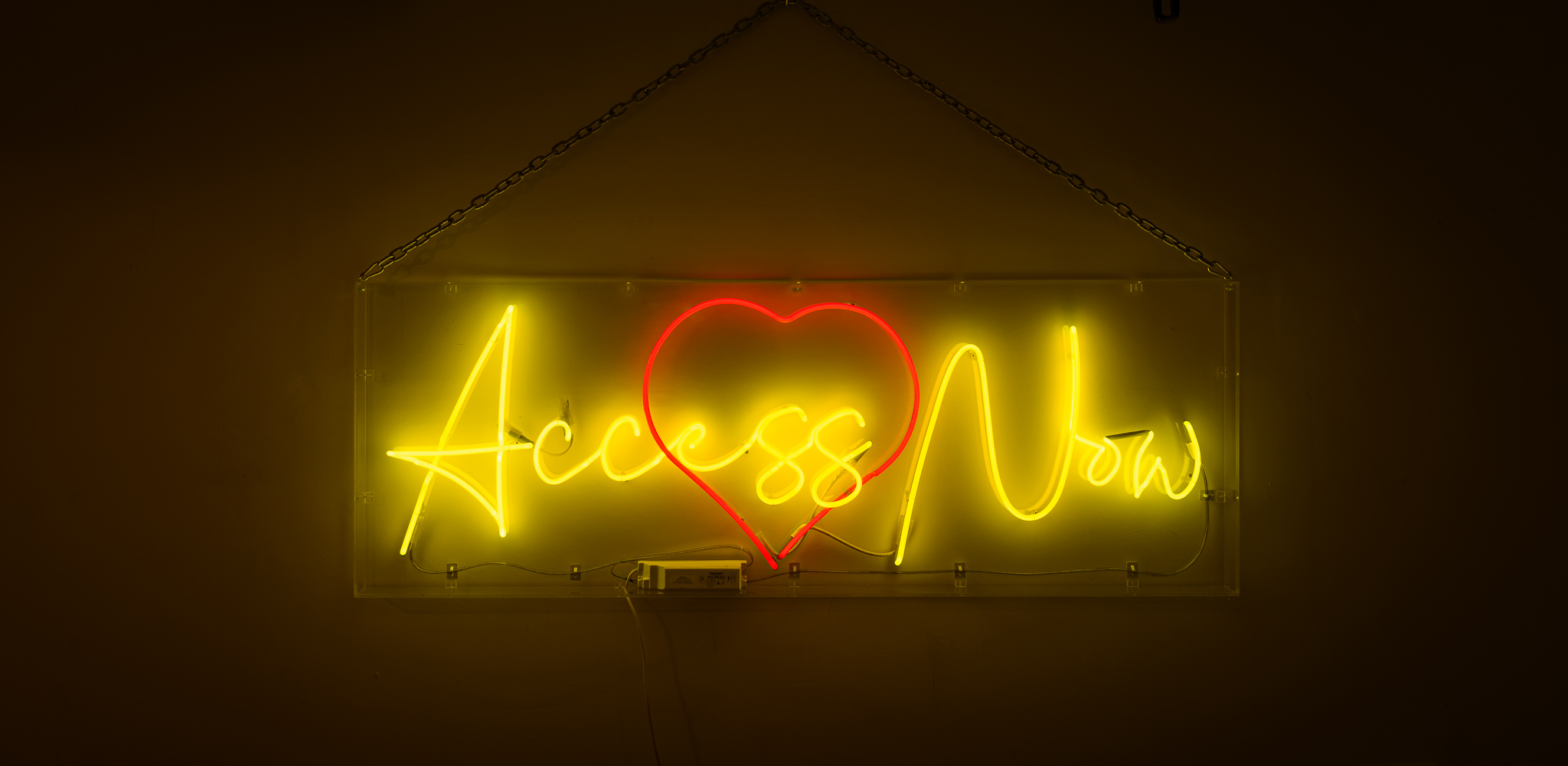 The wording reads on the artwork, 'Access Now'- bright yellow wording with a love heart in red behind the wording, the neon lights are in a stylised handwritten font The neon words are framed inside a transparent perspex box.