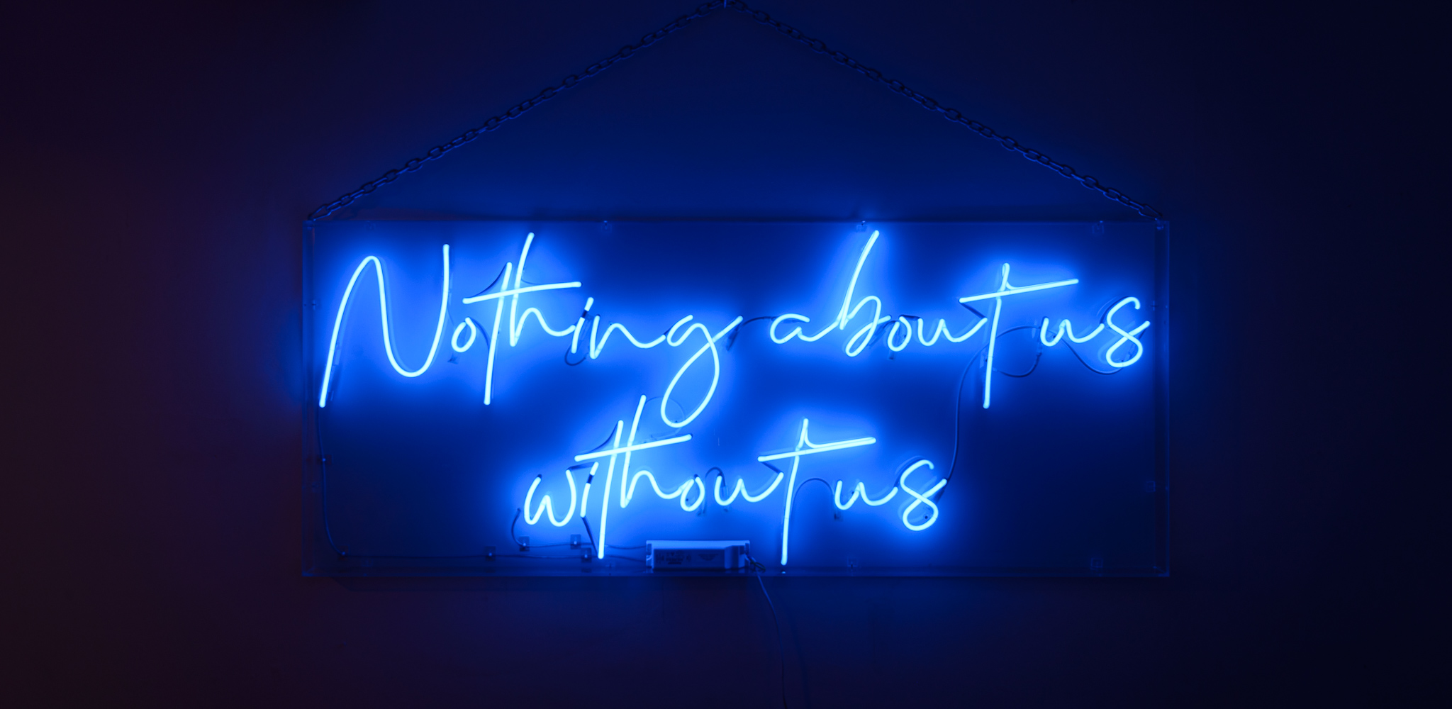 The wording reads on the art work. 'Nothing About Us Without Us',- in dark blue neon lights in a stylised handwritten font The neon words are framed inside a transparent perspex box.