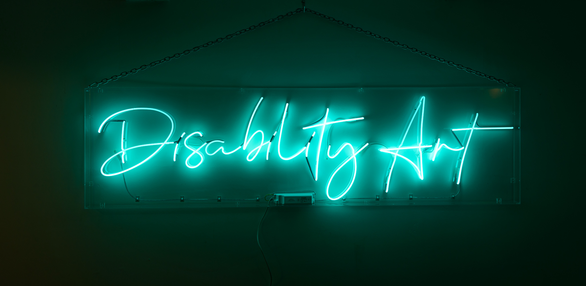 The wording reads on the artwork, 'Disability Art', - turquoise blue neon lights in a stylised handwritten font The neon words are framed inside a transparent perspex box.