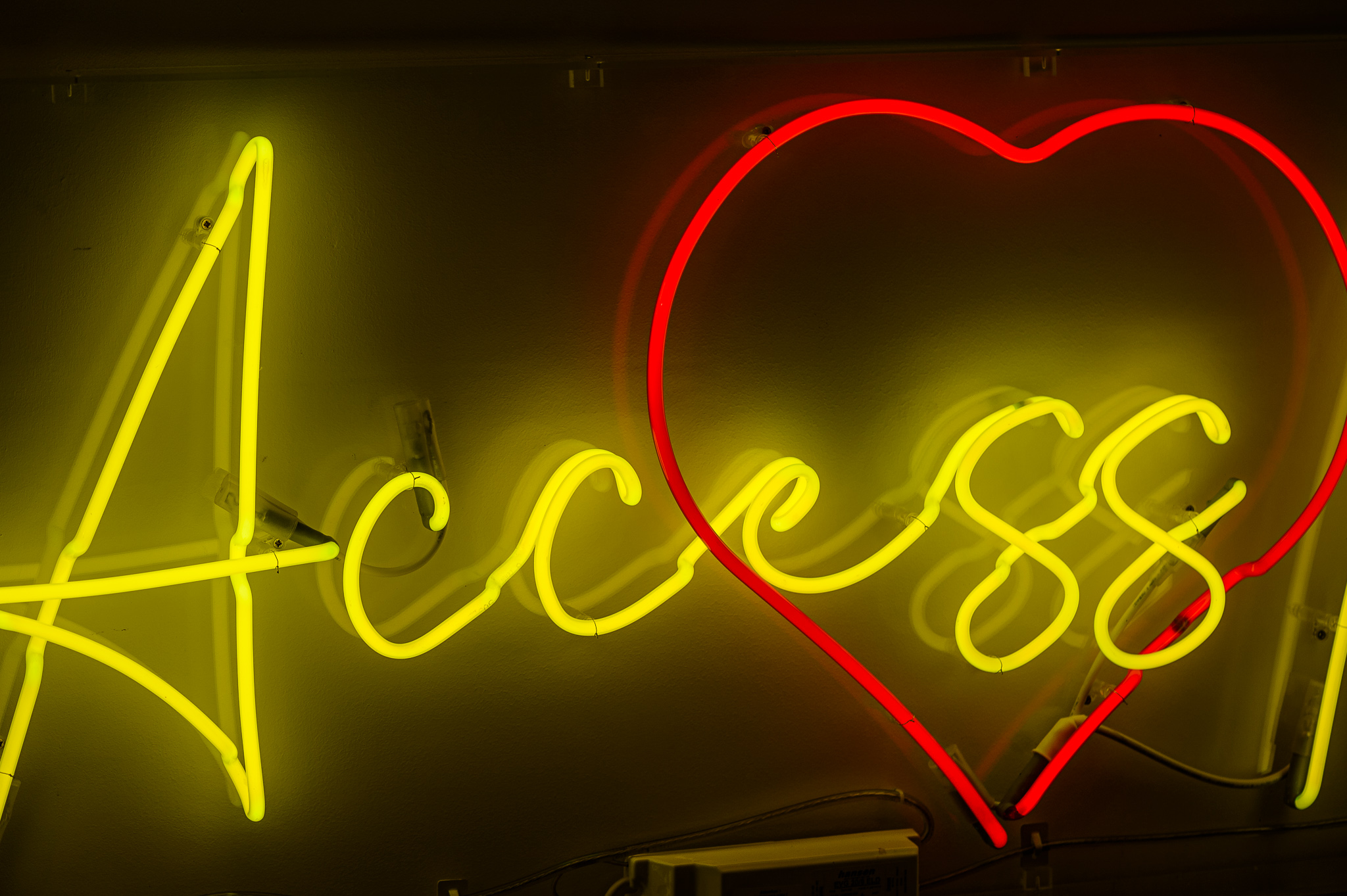 Close up photo of the word 'Access' in bright yellow wording with a love heart in red behind the wording, the neon lights are in a stylised handwritten font The neon words are framed inside a transparent Perspex box.