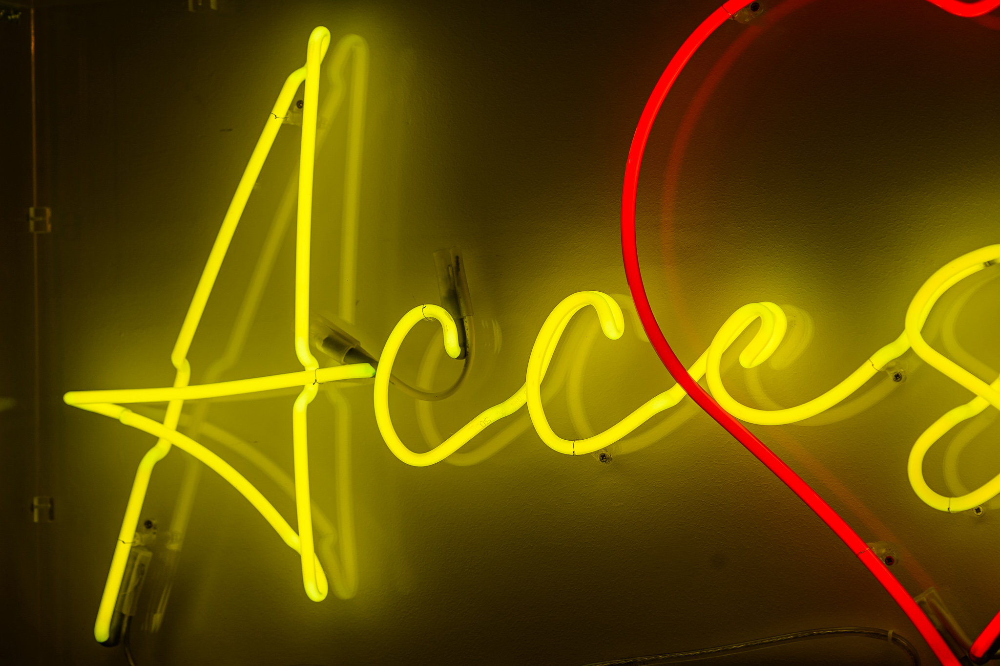 Close up photo of the artwork in bright yellow wording with a love heart in red behind the wording, the neon lights are in a stylised handwritten font The neon words are framed inside a transparent Perspex box.