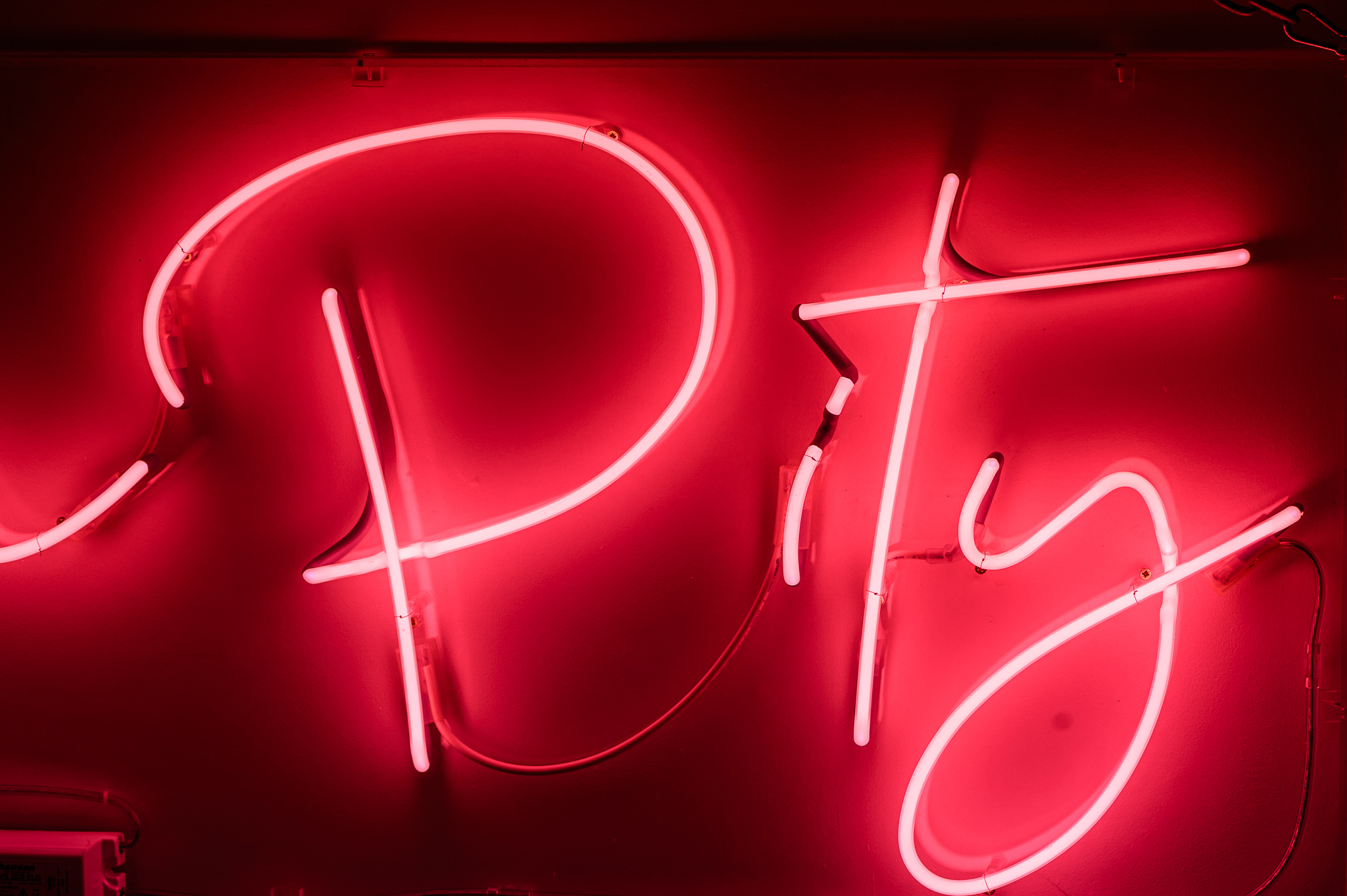 Close up photo on the wird 'Pity' in bright pink neon lights quite flamboyant in a stylised handwritten font. The neon words are framed inside a transparent Perspex box. 