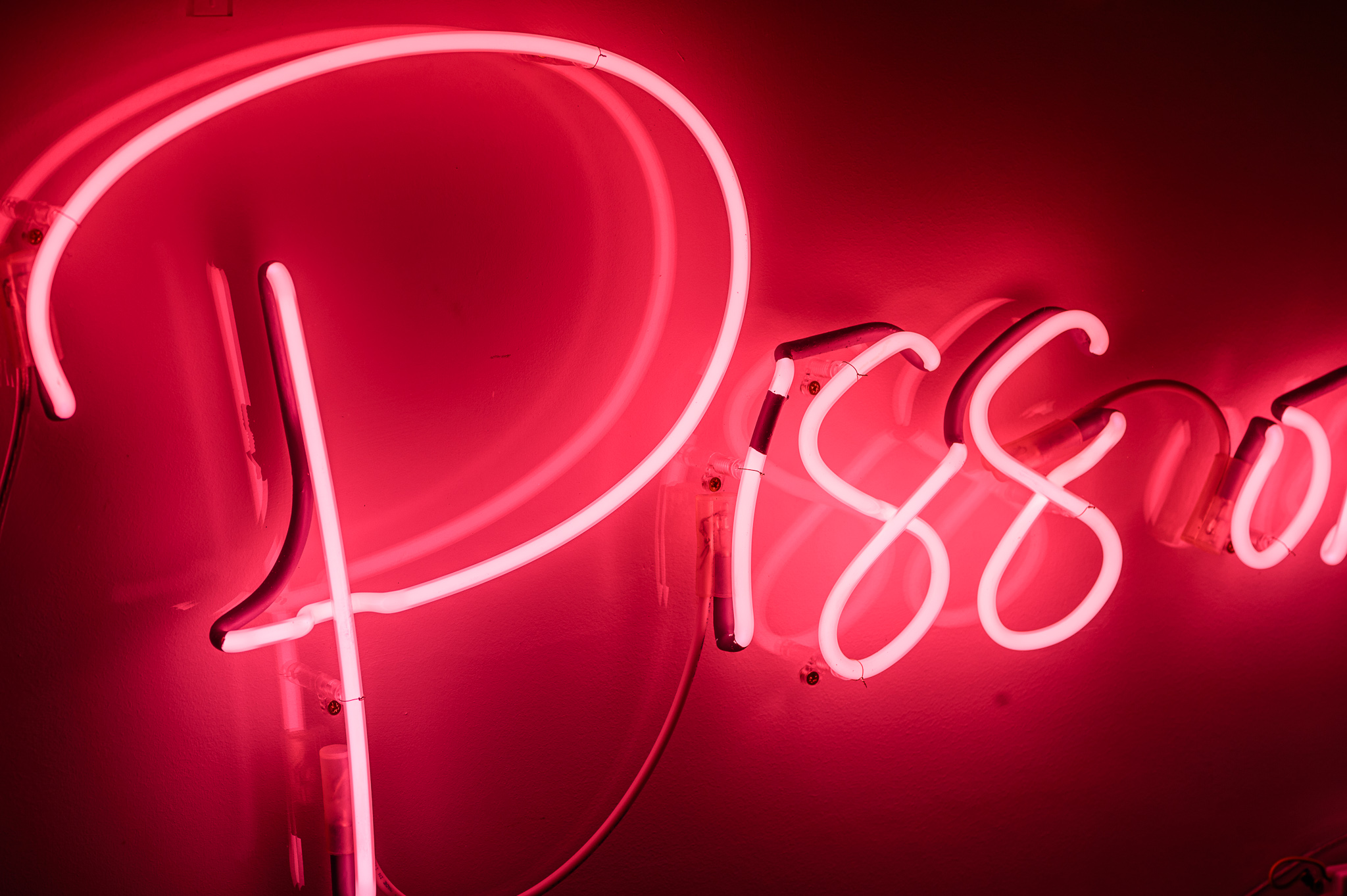 Close up photo on the word 'Piss' in bright pink neon lights quite flamboyant in a stylised handwritten font. The neon words are framed inside a transparent Perspex box. 