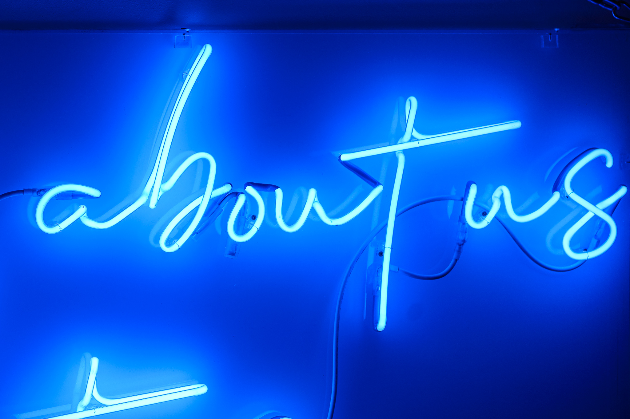 Close up photo of the words 'about us' in dark blue neon lights in a stylised handwritten font The neon words are framed inside a transparent Perspex box. 