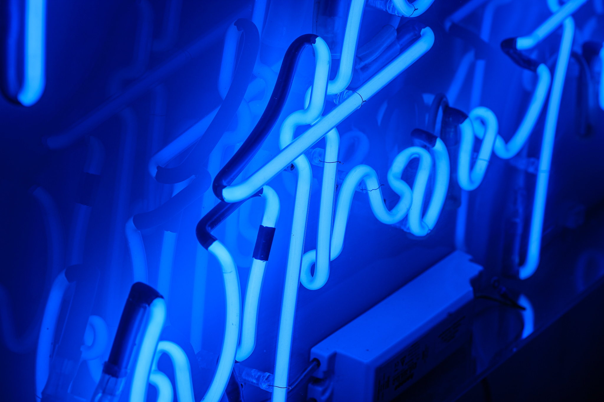 Close up photo of the word 'without' in dark blue neon lights in a stylised handwritten font The neon words are framed inside a transparent Perspex box. 